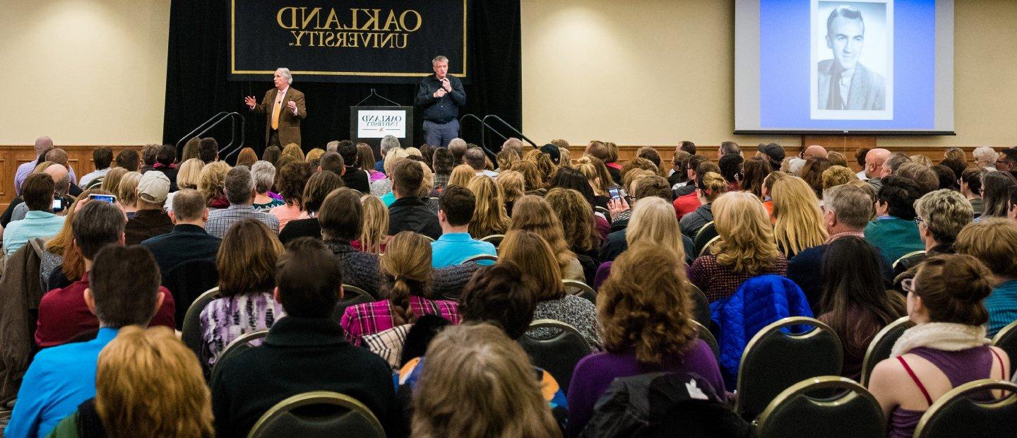 two men standing in front of a black Oakland University banner, addressing a seated audience